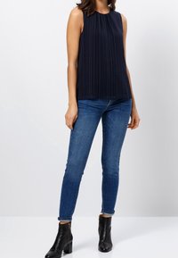 Mörkblå ärmlös blus med vertikala plisseringar, kombinerad med blå skinny jeans och svarta ankelskor med sidodragkedja.