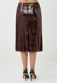 Brauner Pailletten-Midi-Rock mit einem Schlitz auf der Rückseite, der eine glatte Textur und eine figurbetonte Silhouette aufweist. Getragen mit schwarzen High Heels.