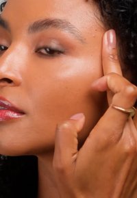 Gros plan d'une personne à la peau lisse pressant doucement le côté de sa tempe avec deux doigts, portant une fine bague en or.