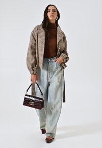 Una modella indossa una giacca over-size a scacchi, un dolcevita marrone, jeans chiari strappati e porta una borsa bordeaux con dettagli dorati.