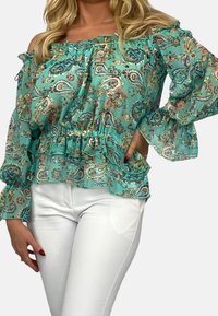 Turkos off-the-shoulderblus med paisleymönster, volangkant och ärmar, tillverkad av lättviktig tyg, tillsammans med vita byxor.