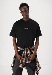 T-shirt preta de algodão com o logótipo "REPLAY" em dourado, combinado com uma camisa de flanela xadrez amarrada, nas cores cinza, branco e laranja.