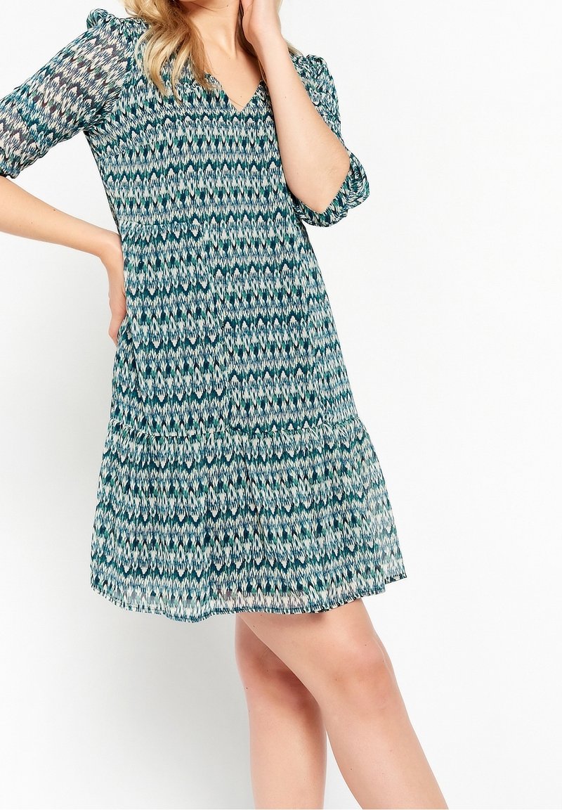 Femme portant une robe longueur genou à motifs bleu, vert et blanc, manches trois-quarts et col en V, posant avec une main sur la hanche.