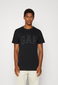 Schwarzes Baumwoll-T-Shirt mit großem, dunkelgrauem "GAP"-Logo auf der Brust. Kurze Ärmel und ein Rundhalsausschnitt runden das Design ab.