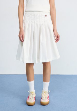White pleated skirt with a waistband, featuring a logo patch. Paired with colorful sneakers and white ribbed socks on a blue floor.