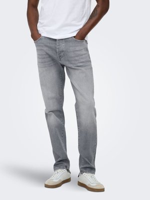 Mann trägt hellgraue Straight-Leg-Jeans, weiße Sneakers und ein weißes T-Shirt, Hände in den Taschen, vor einem einfarbigen Hintergrund stehend.