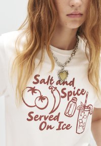 Witte t-shirt met rode grafische tekst en illustraties van tomaten, een chilipeper, een fles en een cocktail, met een zilveren kettinghalsband met een hartbedel.