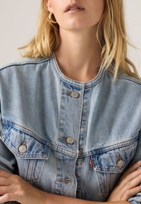 Κοντινό πλάνο ενός ατόμου που φοράει ένα ανοιχτό μπλε τζιν μπουφάν χωρίς γιακά με ασημένια κουμπιά και χρυσούς κρίκους σκουλαρίκια, με τα χέρια σταυρωμένα.