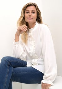 White blouse with ruffled front and cuffs, paired with blue jeans. Smooth fabric, long sleeves, and relaxed fit. Minimalistic design.