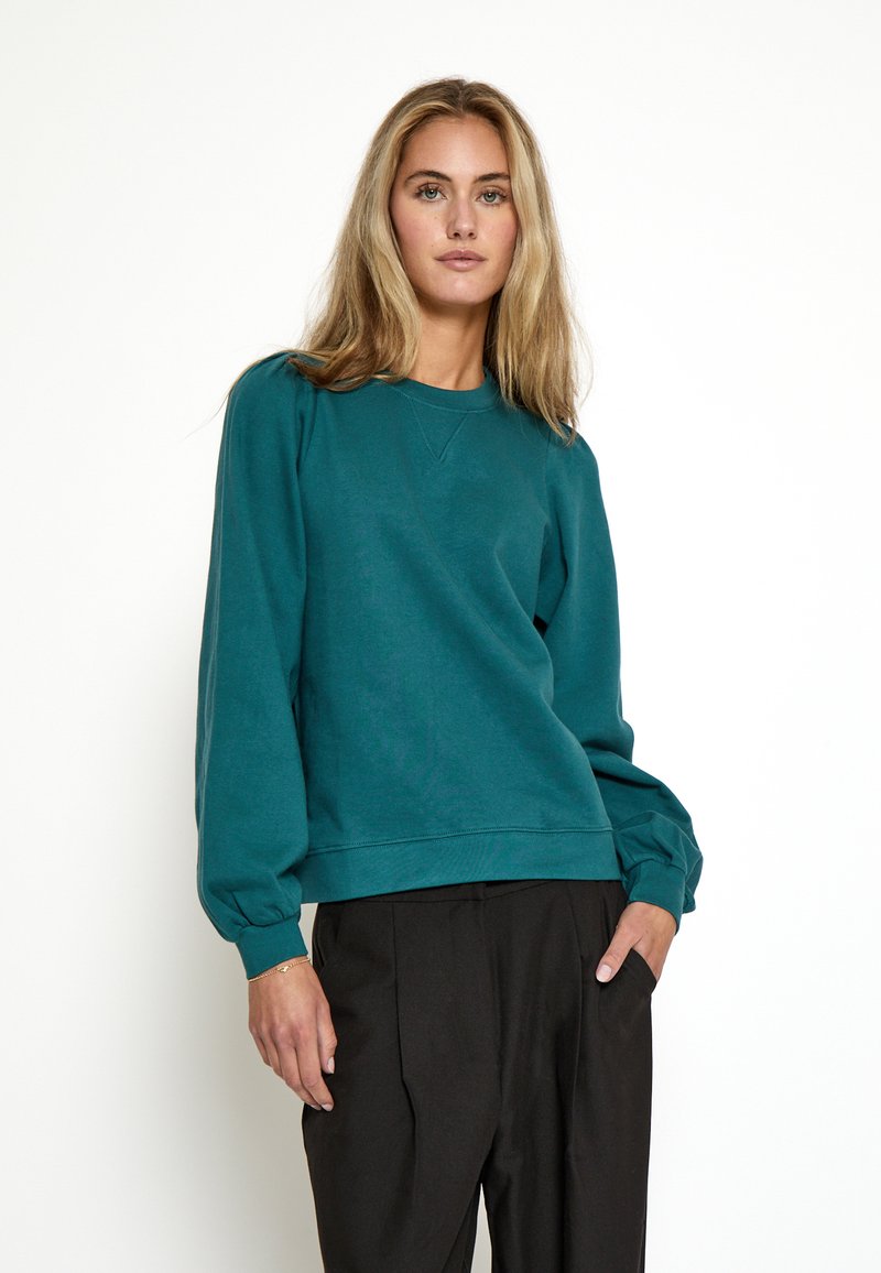 Felpa teal con collo rotondo e maniche a palloncino, caratterizzata da un tessuto morbido e testurizzato e una vestibilità comoda, abbinata a pantaloni neri.
