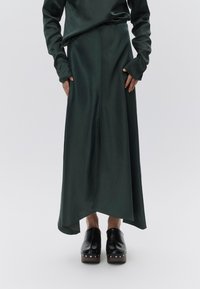 Μακριά πράσινη σατέν φούστα με ασύμμετρο τελείωμα, συνοδευόμενη από ένα ταιριαστό πράσινο τοπ. Μαύρα πλατφόρμα παπούτσια με μεταλλικά τρουκς στη  βάση.