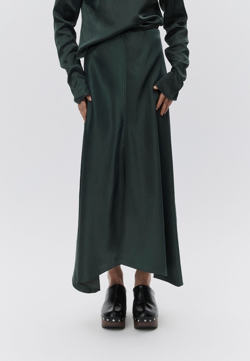 Μακριά πράσινη σατέν φούστα με ασύμμετρο τελείωμα, συνοδευόμενη από ένα ταιριαστό πράσινο τοπ. Μαύρα πλατφόρμα παπούτσια με μεταλλικά τρουκς στη βάση.