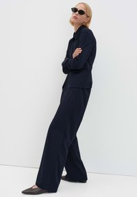 Marineblauw op maat gemaakt overhemd en broek met wijde pijpen, gemaakt van gladde stof. Accessoires omvatten zwarte ronde zonnebril en puntige donkere schoenen.