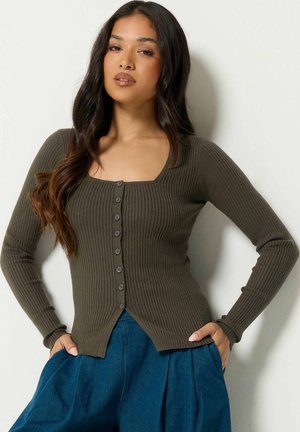 Femme aux longs cheveux foncés portant un cardigan côtelé boutonné vert olive et un pantalon bleu plissé taille haute, debout les mains dans les poches.