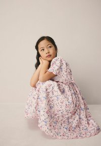 Mlada deklica v rožnati obleki sedi na tleh, z rokami počiva na obrazu in premišljeno gleda vstran.