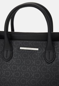 Sac à main noir Calvin Klein avec doubles poignées, surface texturée présentant un motif répété "CK" et plaque logo de la marque en argent.