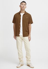 Camisa marrón de mangas cortas con cierre de botones y bolsillo, llevada sobre una camiseta blanca. Combinada con pantalones beige claros y zapatillas blancas con detalles marrones.