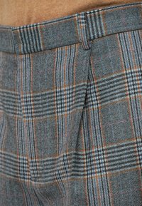 Pantalons à carreaux gris avec une taille structurée et un mélange de lignes bleues, noires et oranges, en tissu texturé et avec de légères plis.