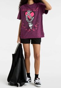 Camiseta gráfica morada con un diseño de corazón y cadena, combinada con pantalones cortos negros y una chaqueta de béisbol negra, sostenida en la mano.
