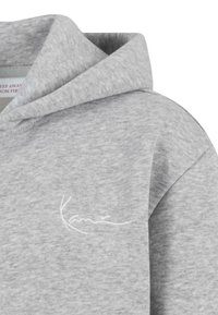 Světlé šedé mikina s kapucí s bílým vyšitým logem na hrudi, vyrobená z měkké texturované látky.