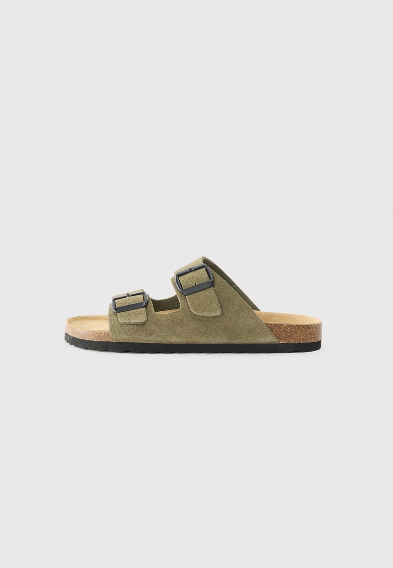 Sandales en daim vert avec deux boucles réglables, semelle en liège et semelle extérieure texturée noire. Design à bout ouvert, silhouette plate.