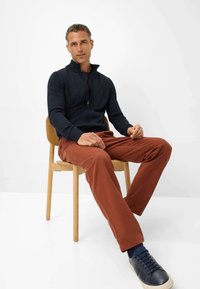 Marineblå lynlåstrøje, rustfarvede bukser og marineblå sneakers. Model siddende på en træstol med en enkel hvid baggrund.