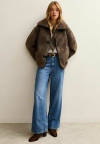 Kobieta w brązowej puszystej kurtce, swetrze w romby, niebieskich dżinsach z szerokimi nogawkami i brązowych botkach na obcasie stoi przy beżowej ścianie.