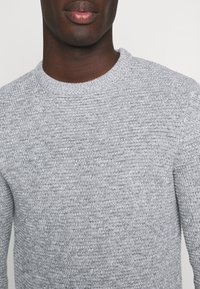 Grauer Strickpullover mit rundem Ausschnitt, der ein strukturiertes Muster und gerippte Bündchen aufweist. Der Stoff wirkt weich und gemütlich.