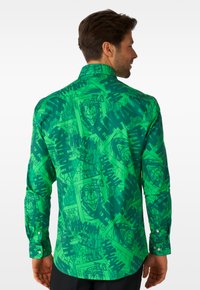 Ein Mann von hinten, der ein grünes Langarmshirt trägt, bedruckt mit Joker-Spielkarten und sich wiederholenden "HAHA"-Textmustern.