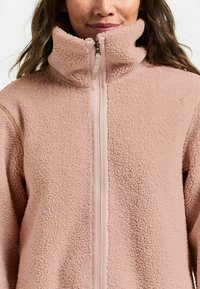 Veste en polaire rose avec un col montant et une fermeture éclair frontale, présentant une surface texturée et une bordure en tissu lisse le long de la fermeture éclair.