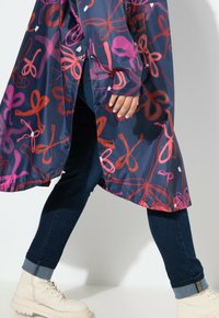 Giacca navy resistente all'acqua con motivi astratti rosa e rossi, bottoni a pressione e un orlo più lungo, abbinata a jeans scuri e stivali beige.