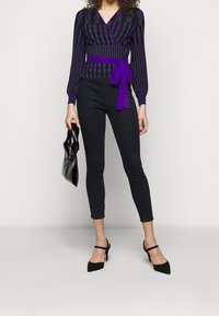 Fialovo-černý pruhovaný zavinovací top s tkaničkou v pase, ve spojení s černými přiléhavými kalhotami a špičatými černými lodičkami, držící černou kabelku.