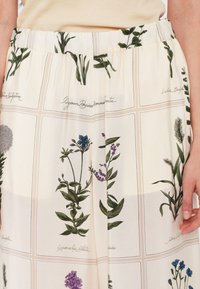 Pantalon ample de couleur crème avec imprimé botanique présentant différentes plantes vertes et des noms latins manuscrits en motif quadrillé.