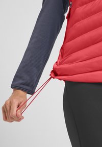 Gilet rosso imbottito con design trapuntato e cordoncino in vita, indossato sopra una maglietta a maniche lunghe blu scuro. Finitura lucida e zip a contrasto.