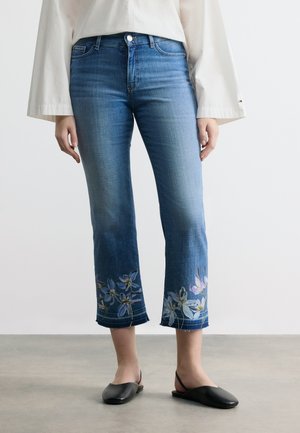 Kobieta ubrana w niebieskie krótkie jeansy z kwiatowym haftem przy nogawkach, w połączeniu z czarnymi butami wsuwanymi i białą bluzką z długim rękawem.