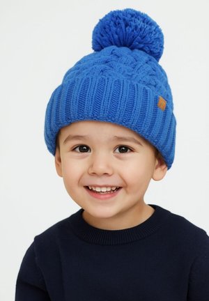 Modra pletena kapa z pompomom na vrhu, ki ima strukturo in majhno rjavo usnjeno oznako, jo nosi otrok v temni majici.