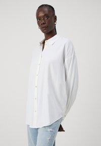 White long-sleeve shirt with a buttoned collar, featuring a loose fit and soft texture. Paired with light blue distressed denim jeans.