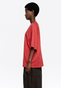 Camiseta roja oversized con mangas cortas y cuello redondo. Material de algodón suave. Modelo lleva una falda oscura de tiro alto. Vista de perfil lateral.