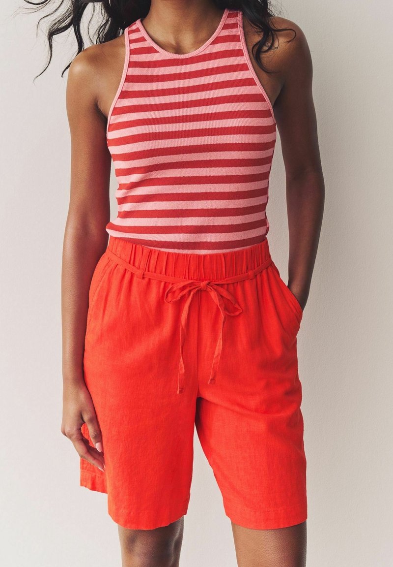 Femme portant un haut sans manches rayé rouge et rose et un short à cordon rouge vif, debout devant un fond clair uni.