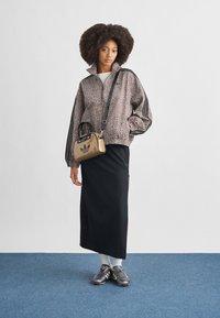 Ανοιχτό καφέ ζακέτα με φερμουάρ και σχέδια, με μαύρες ρίγες, σε συνδυασμό με μια μαύρη μακριά φούστα, ασημένια παπούτσια και μια μικρή μπεζ τσάντα με λογότυπο.