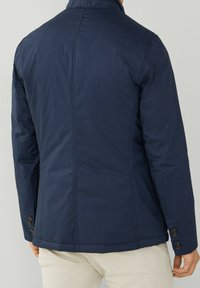 Marinblå, lätt jacka med ståkrage, knappdetaljer vid ärmslut och sömaccenter. Texturerad tyg med modern passform.