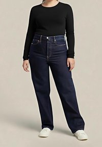 Personne portant un haut noir moulant à manches longues, un jean bleu foncé taille haute et des chaussures blanches sans lacets, debout devant un fond uni.