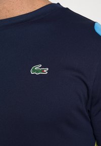 Tmavě modrá krátký rukáv košile s texturovanou tkaninou, s malým zeleným krokodýlím logem na hrudi a modrými akcenty na ramenech.