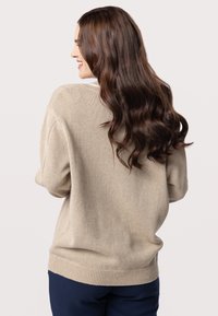 Maglione beige lavorato a maglia con una vestibilità rilassata, polsini e orlo a coste, caratterizzato da una texture sottile. Il modello ha i lunghi capelli che cadono sulla spalla.