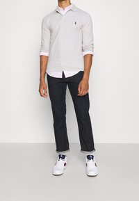 Man som bär en ljusgrå långärmad polo över en rosa randig skjorta, mörka uppvikta jeans och vita sneakers med marinblå och röda detaljer.
