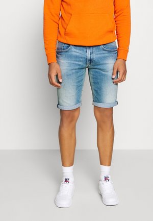 Mężczyzna noszący podwinięte jasnoniebieskie denimowe szorty, pomarańczową bluzę, białe skarpetki Reebok i białe trampki, stojący na jednolitym tle.