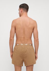 Hnedé plážové šortky vyrobené z ľahkého materiálu, s elastickým pásom a bielymi akcentmi loga Adidas, majú jednoduchý dizajn.