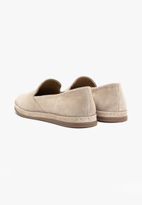 Beżowe zamszowe buty wsuwane z zaokrąglonymi czubkami, niską, płaską brązową podeszwą i widocznym przeszyciem, przedstawione z tyłu pod kątem trzech czwartych.