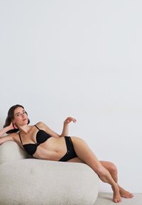 Černá lingerie souprava s vyztuženou podprsenkou a kalhotkami ve stylu bikiny. Měkká tkanina, minimalistický design a jemné ramínka. Umístěná na texturované světlé pohovce.