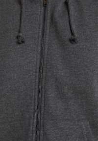 Sudadera gris con cremallera de tejido suave, con capucha ajustable mediante cordón, que presenta una cremallera frontal y dos bolsillos laterales. La textura parece suave.